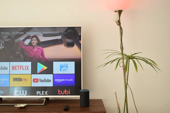 Amazon Alexa accende una lampadina. Dietro una televisione che mostra l'interfaccia di Amazon Fire TV
